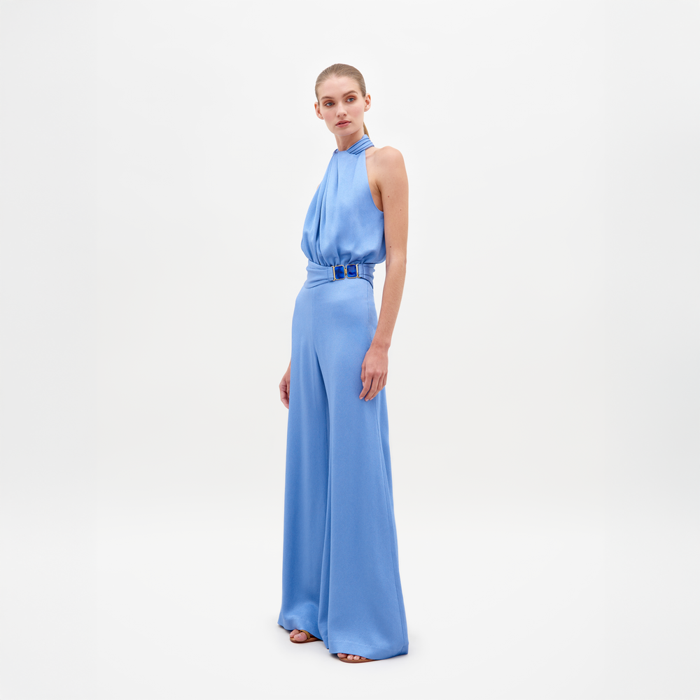 Con el mono Siv, una persona posa sobre un fondo blanco. El elegante mono azul claro presenta un escote halter y pantalones anchos de satén martillado con cintura entallada, que acentúa bellamente su silueta mientras mira ligeramente hacia un lado.