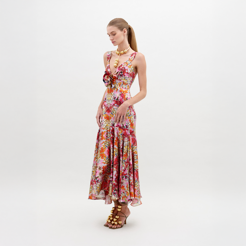 Una mujer con el Vestido Saskya se encuentra de pie frente a un fondo blanco. Este colorido vestido floral midi presenta un diseño de apliques en 3D con un escote con volantes y un dobladillo acampanado. Lo complementa con tacones adornados con flores, un collar dorado y el cabello peinado en una elegante cola de caballo.
