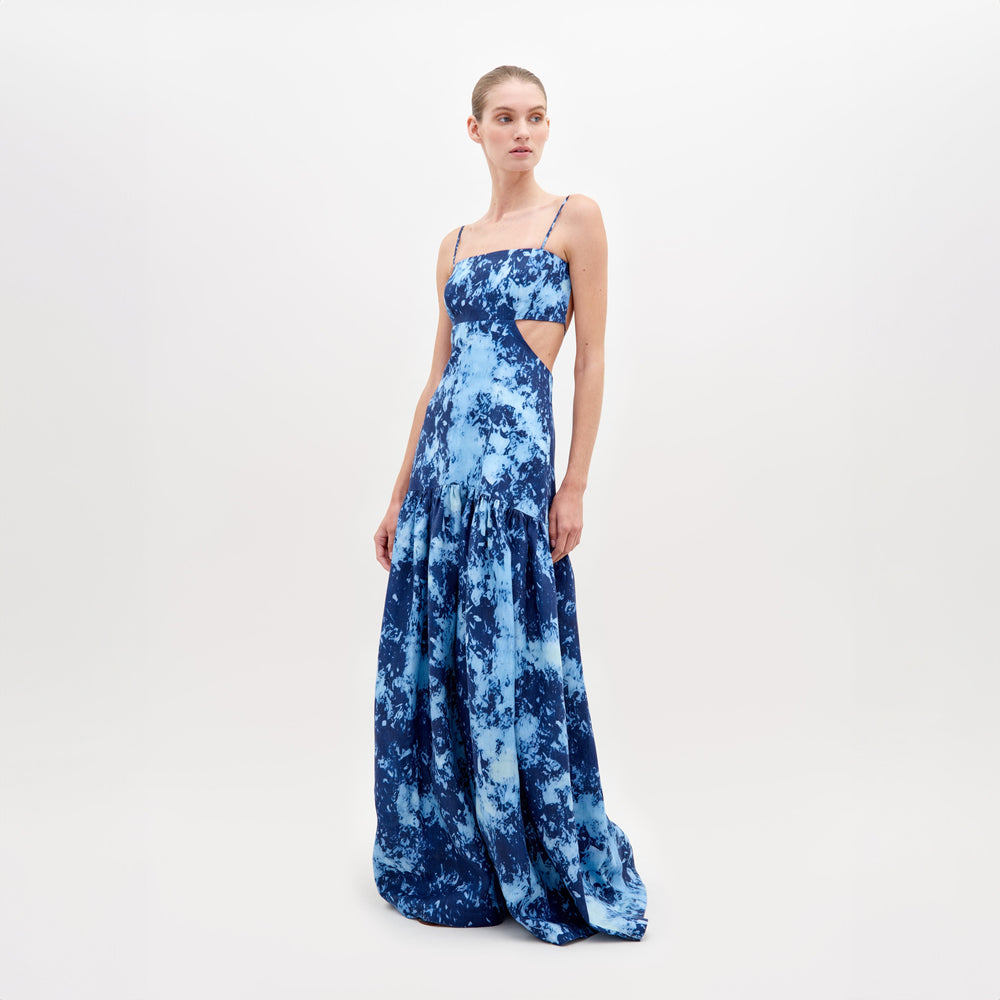 Una mujer posa con el Vestido Shannon, un vestido largo azul con efecto tie-dye, tirantes finos y aberturas en los costados. Presenta un corpiño entallado que fluye hacia una falda amplia. Ella está de pie sobre un fondo blanco liso.