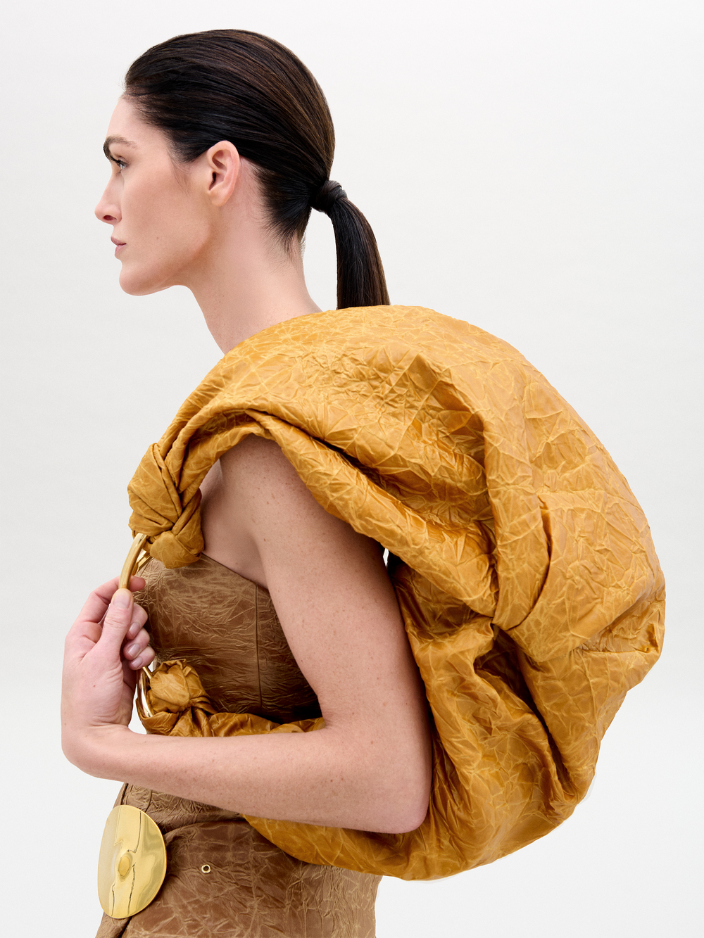Una persona de cabello oscuro con una elegante cola de caballo luce un conjunto marrón texturizado de tela italiana, realzado por un drapeado mostaza sobre el hombro. El conjunto se complementa con un cinturón ancho con hebilla redonda dorada y un elegante bolso Sofia Maxi.