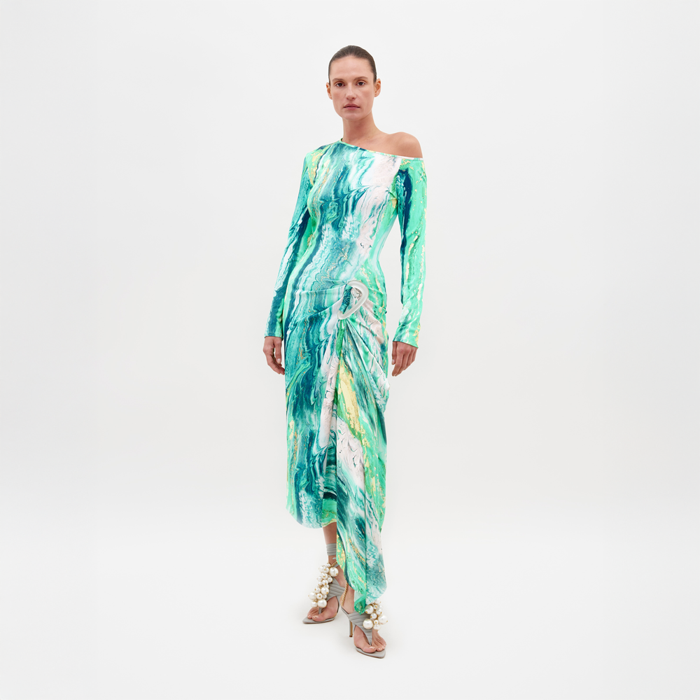 Sobre un fondo blanco liso, una persona luce el Vestido Tahia, un vestido largo de un solo hombro con estampados abstractos en verde y azul. Lo combina con sandalias adornadas con grandes adornos en forma de conchas.
