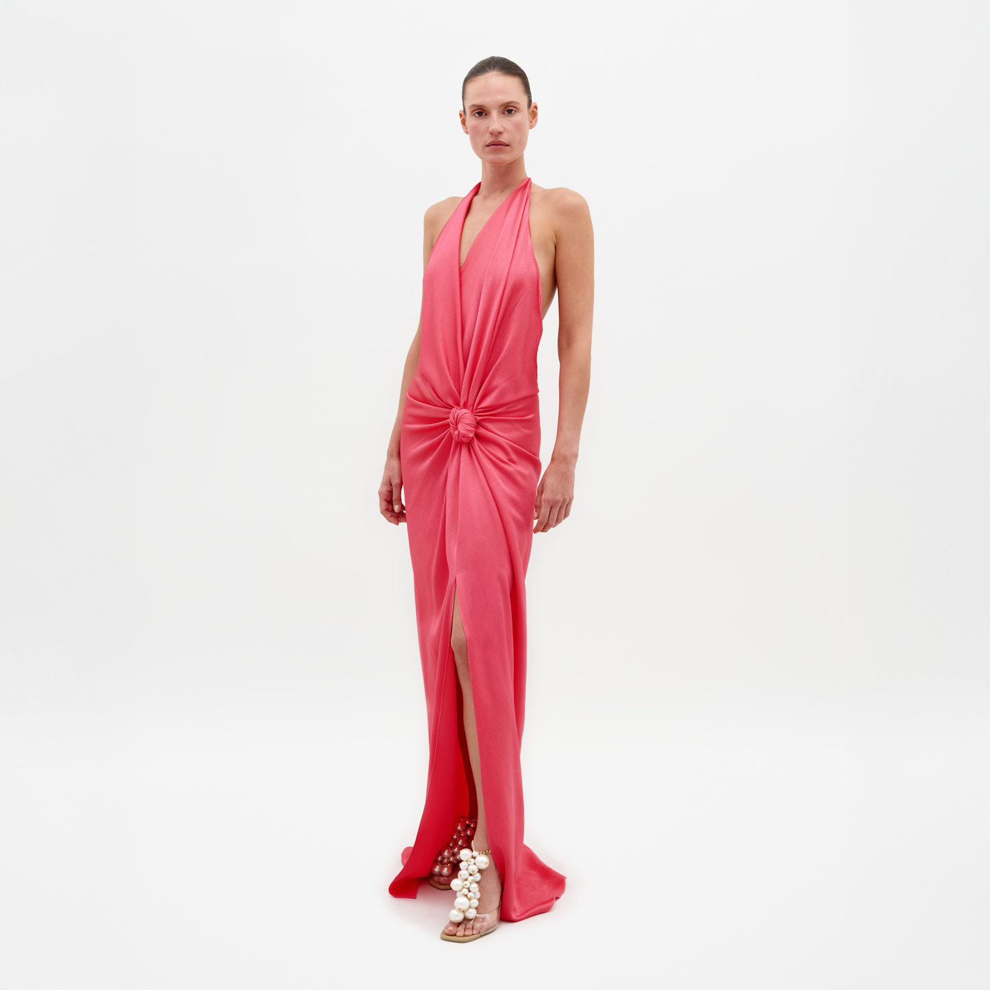 Una persona lleva el Vestido Torgiano rosa sin mangas, un vestido largo con cuello halter, detalle de nudo en la cintura, abertura delantera y espalda abierta. Está combinado con sandalias con adornos esféricos sobre un fondo blanco liso.