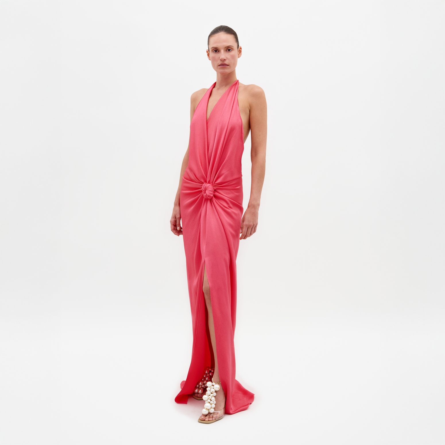 Una persona lleva el Vestido Torgiano rosa sin mangas, un vestido largo con cuello halter, detalle de nudo en la cintura, abertura delantera y espalda abierta. Está combinado con sandalias con adornos esféricos sobre un fondo blanco liso.