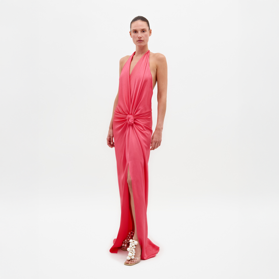 Una persona lleva el Vestido Torgiano rosa sin mangas, un vestido largo con cuello halter, detalle de nudo en la cintura, abertura delantera y espalda abierta. Está combinado con sandalias con adornos esféricos sobre un fondo blanco liso.