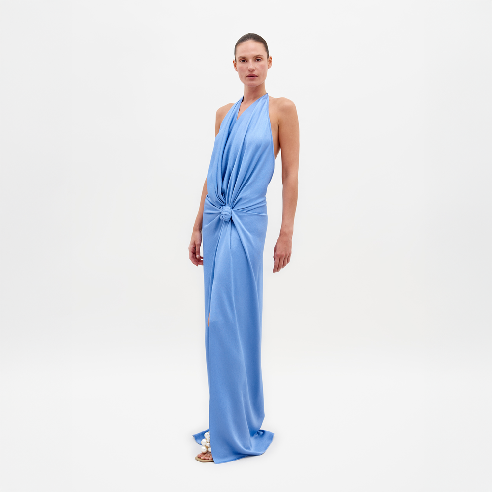 Una persona está de pie descalza, luciendo el fluido Vestido Torgiano azul con cuello halter y un gran detalle de nudo en la cintura. Sobre un fondo blanco liso, esta exquisita pieza está orgullosamente confeccionada en Colombia.