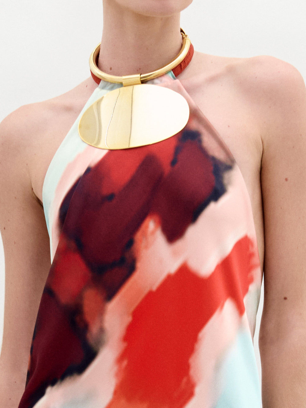 Una persona viste el Vestido Zeffa, un vestido largo con escote halter que presenta un patrón de acuarela intenso en rojo, negro y blanco. Un collar de oro sustancial con un gran colgante ovalado adorna su cuello. El elegante conjunto, hecho en Colombia, se destaca contra el fondo blanco liso.