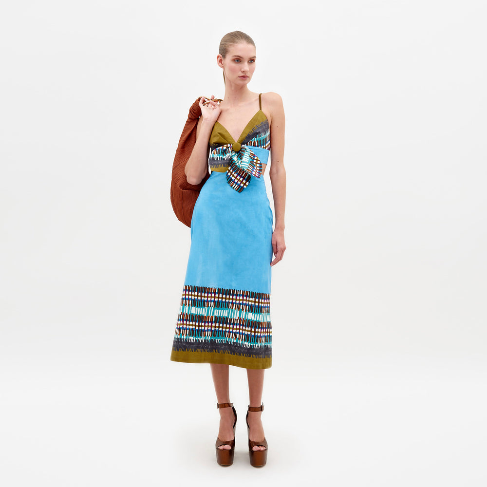 A woman is wearing the Atira Skirt in an aqua olive plaid pattern along with a large bow. She has an orange jacket draped over her shoulder and is standing against a plain white background. This look will be available for pre-order starting February 15th, 2025.